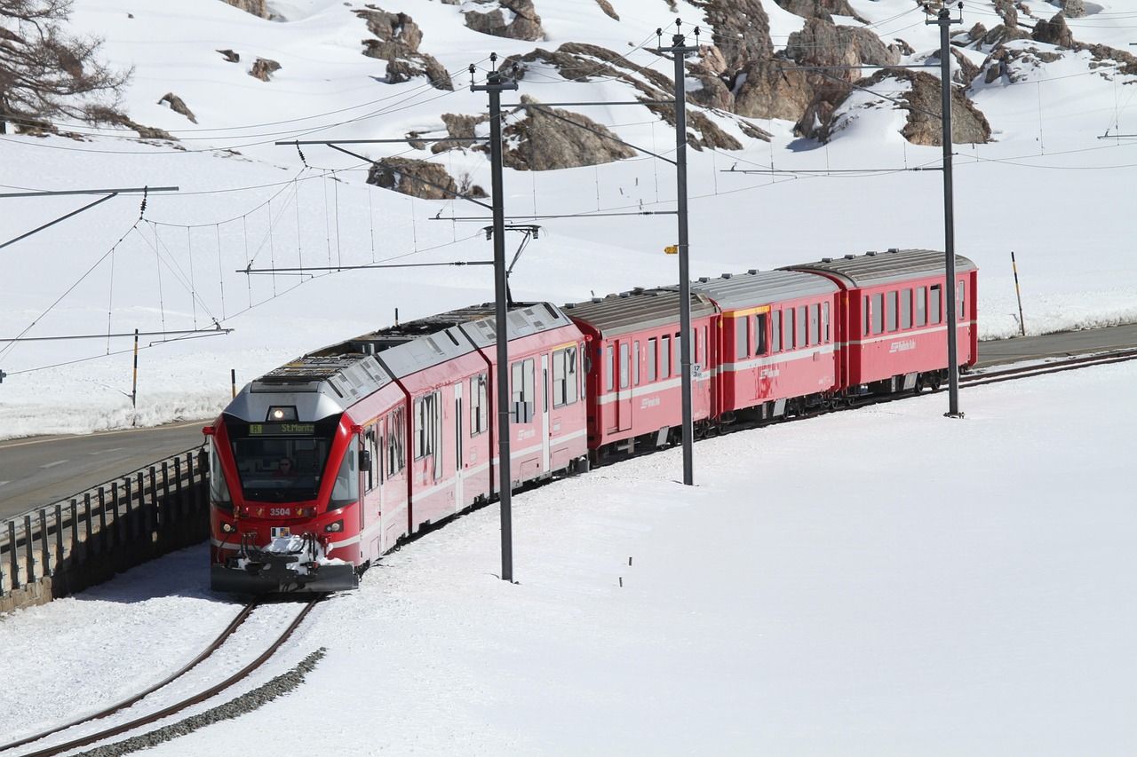 Trenino rosso che viaggia sui binari in un paesaggio innevato, con le montagne sullo sfondo.