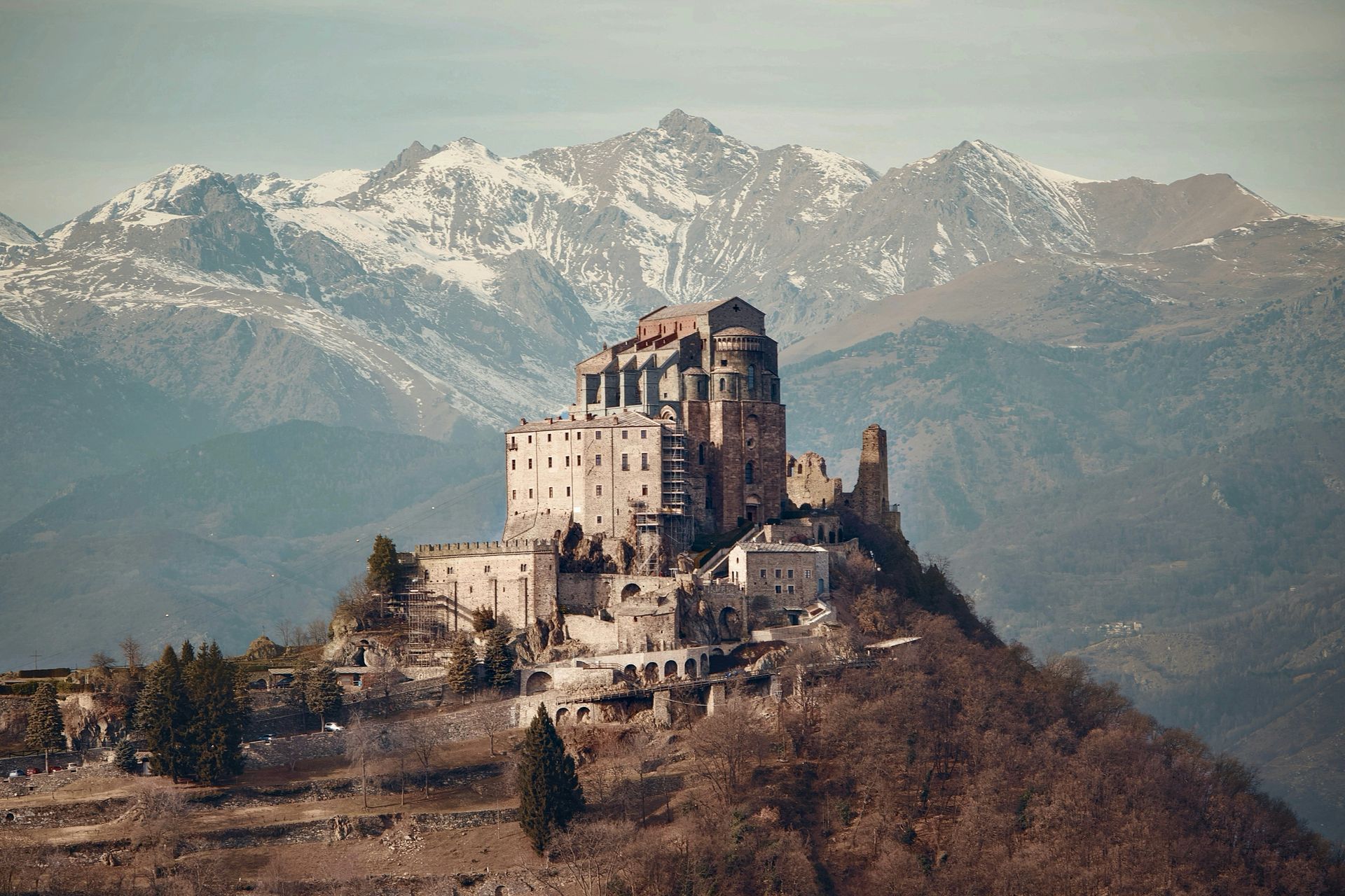 Monastero in pietra arroccato sulla cima di una collina con montagne innevate sullo sfondo.
