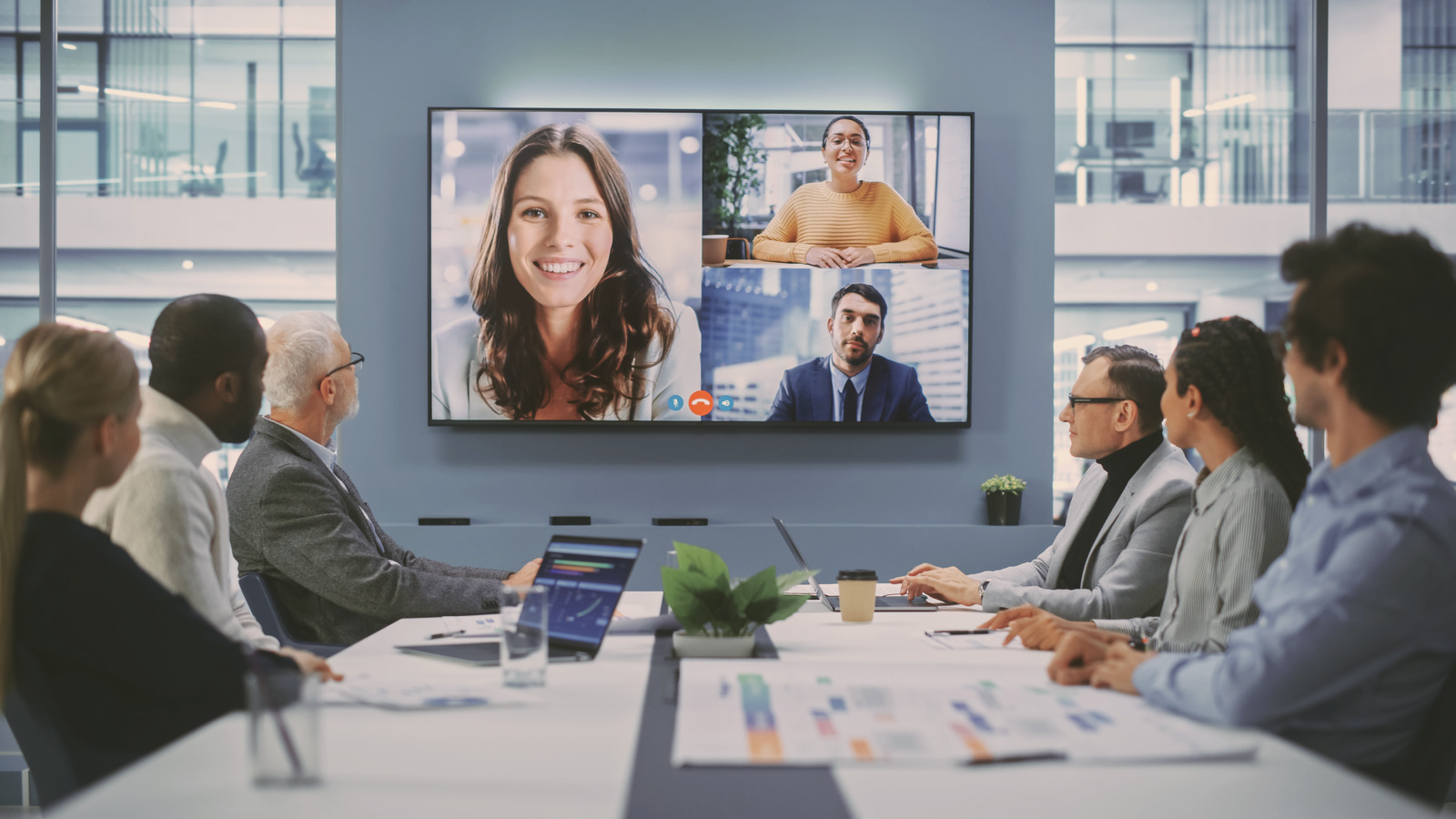 Business meeting in a glass-walled conference room with a large screen showing a video call with three participants.
