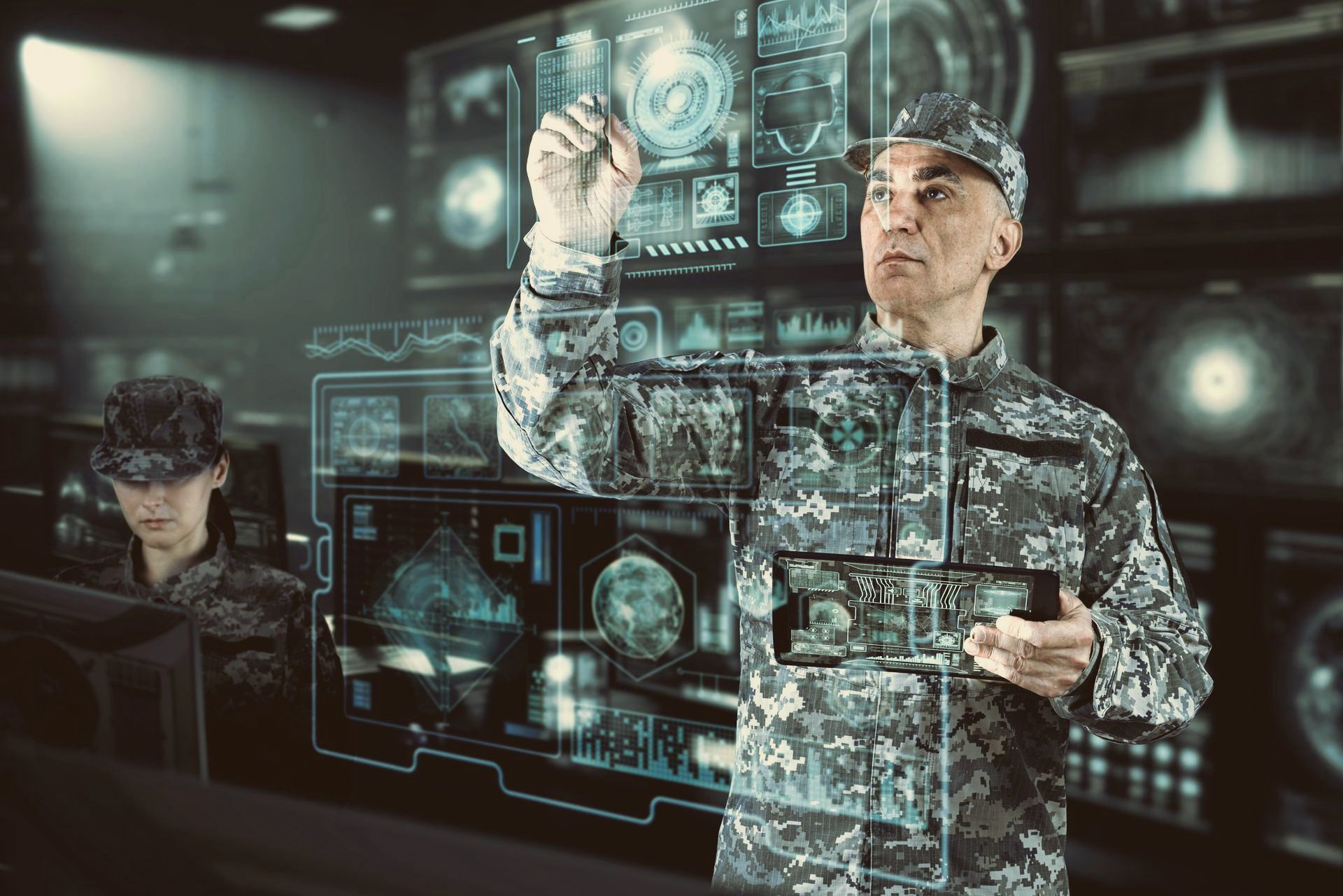 Two people in camouflage operate a transparent digital control interface in a dark command center