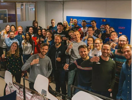 Group of smiling people posing indoors at an event, holding drinks in front of a blue backdrop.