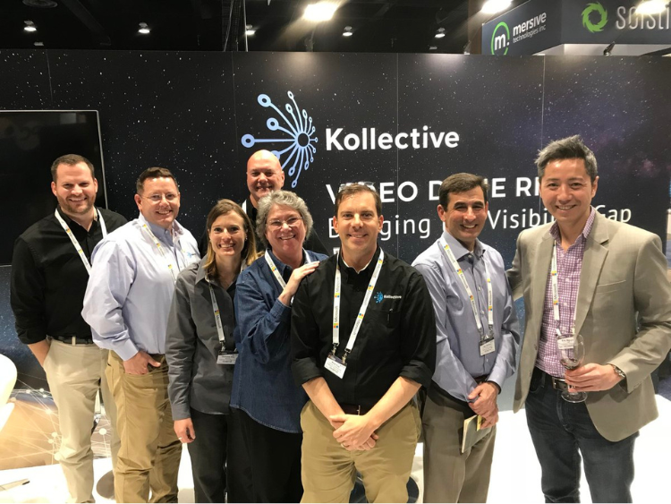 Seven people posing at a trade show booth with a black Kollective backdrop and smiling faces
