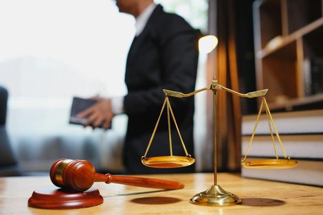 Gavel and balance scale on a desk, with a blurred lawyer holding a brief in the background.