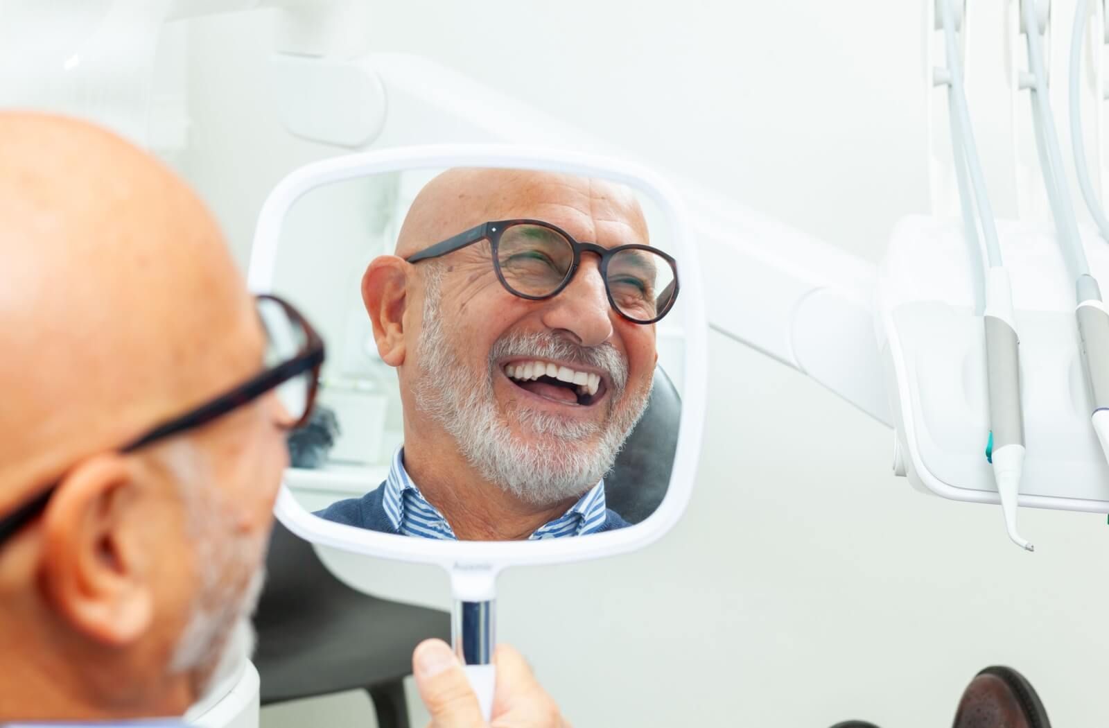 A reflection in a handheld mirror of a smiling senior man with a beard and glasses, admiring his healthy dental implants while at the dentist’s office.