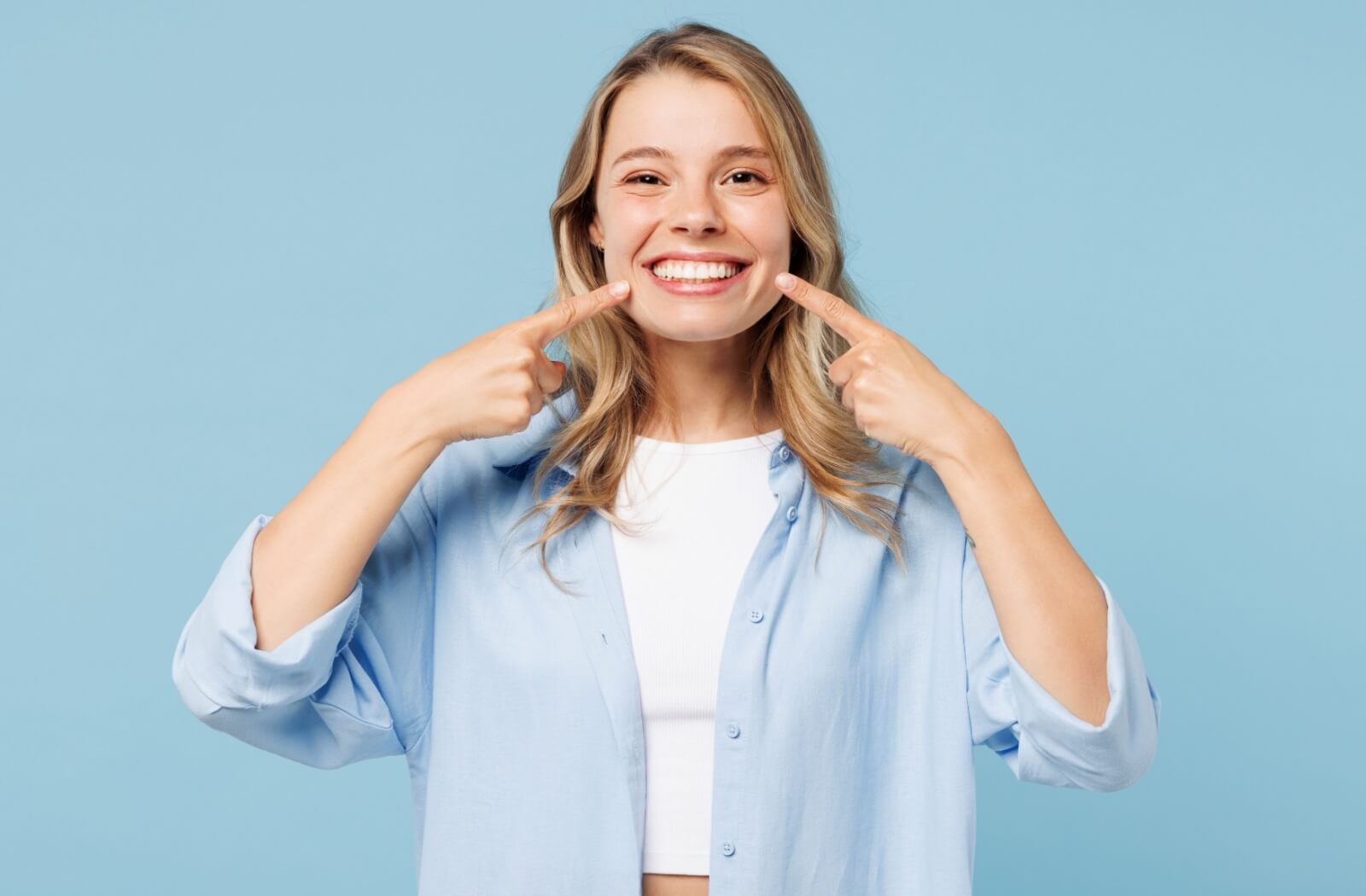 A happy woman in a light blue shirt pointing to her perfect, gap-free smile against a solid blue background.