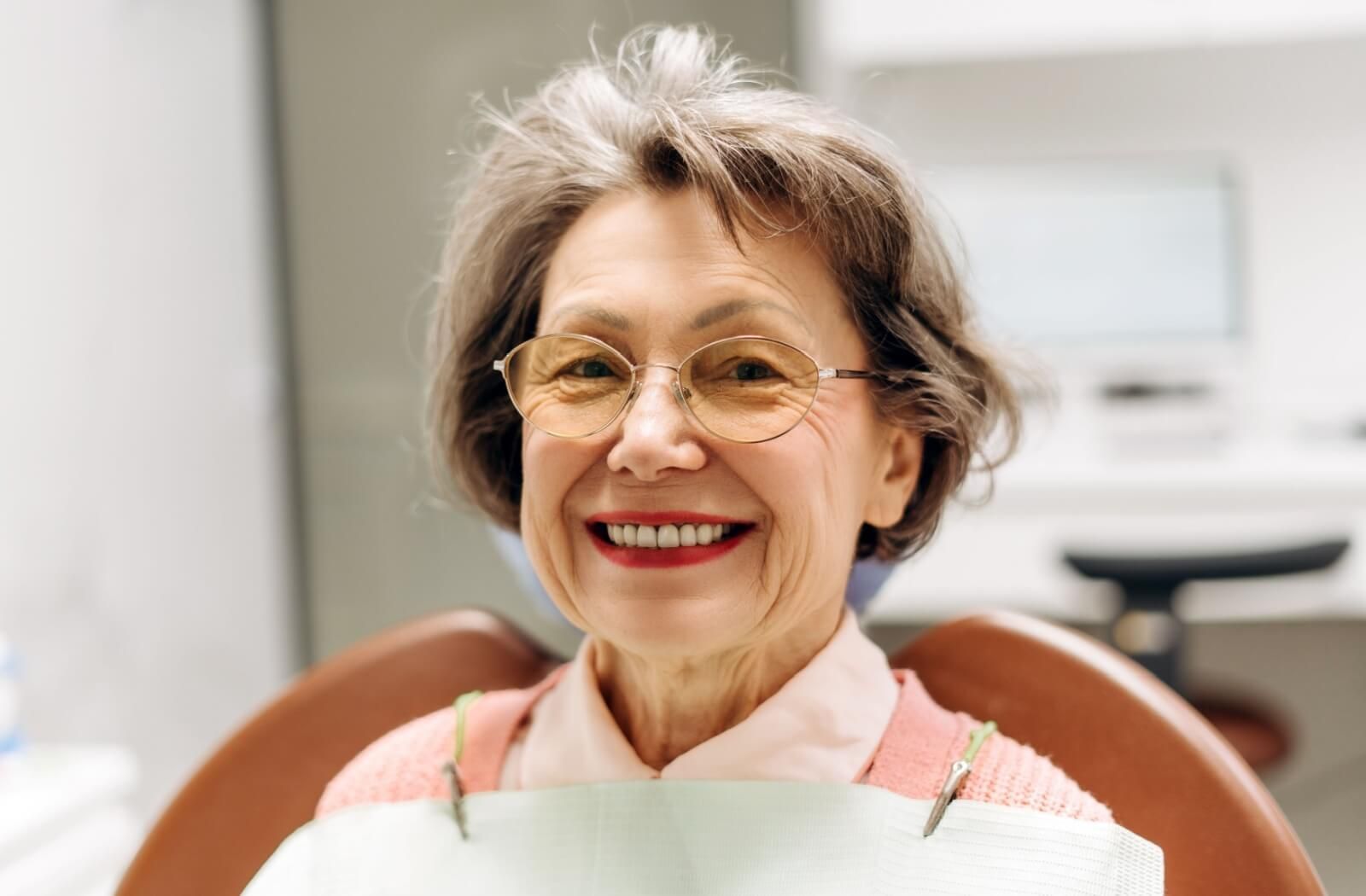 A smiling senior woman with glasses sitting in a dental chair, showcasing a healthy restored smile after a partial denture consultation.
