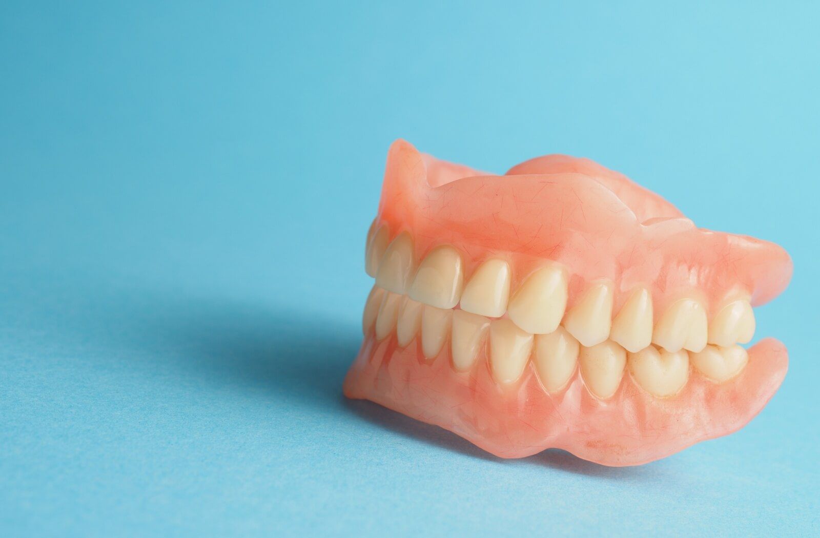 A set of full or partial dentures with pink acrylic gums and white teeth shown against a solid light blue background.