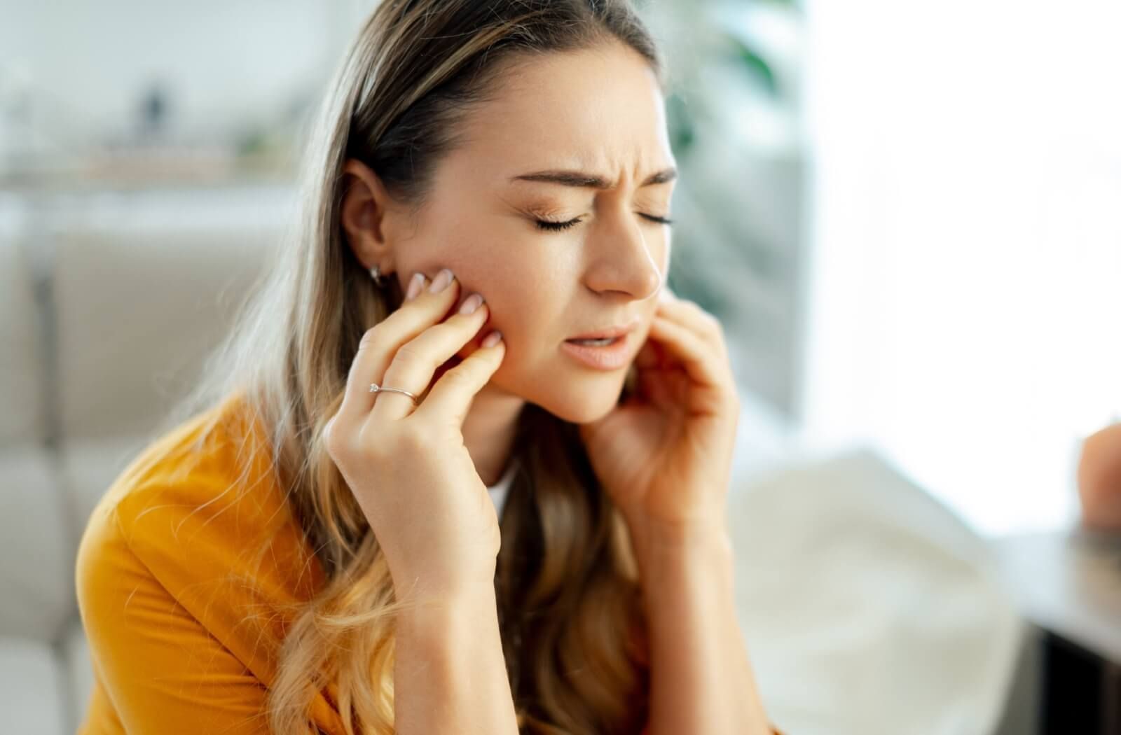 A woman in an orange shirt sitting with her eyes closed and hands pressed against her jaw and temples, illustrating chronic TMJ pain and jaw tension.