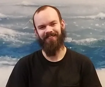 Smiling bearded person in a black shirt standing before a cloudy ocean backdrop