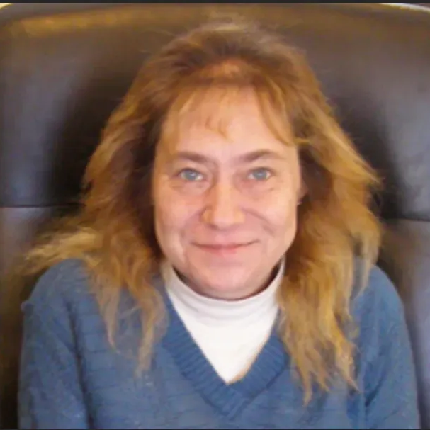 Smiling person in a blue sweater and white turtleneck, seated against a dark leather chair.