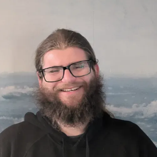 Smiling person with glasses and a black hoodie in front of a cloudy seascape painting