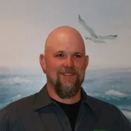 Smiling bald man with a goatee in a dark shirt, standing before a seascape mural with a flying bird.