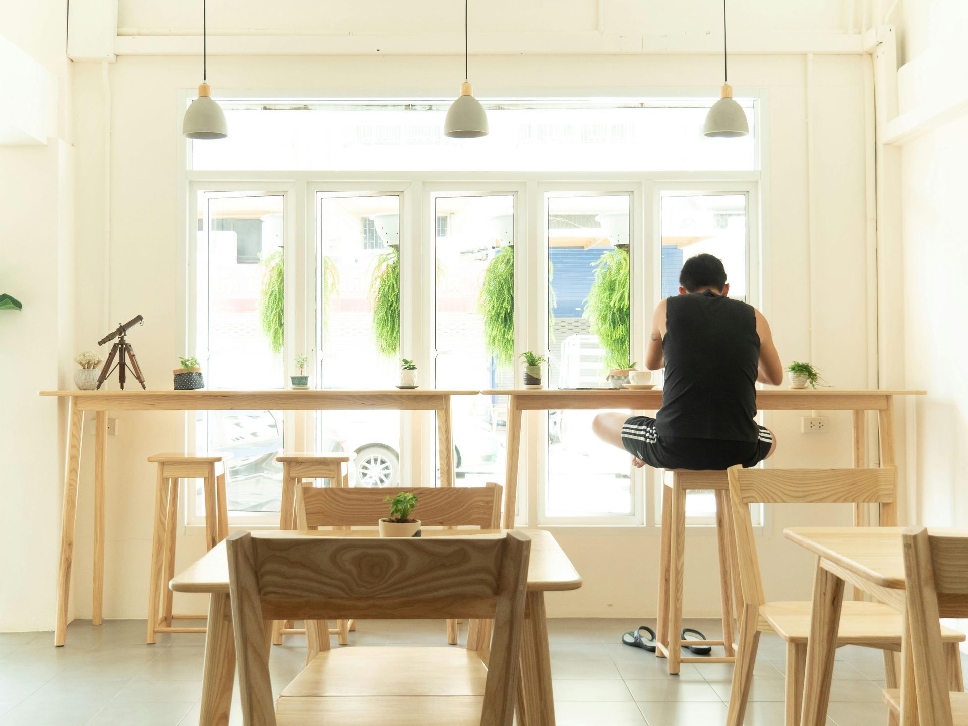 Man sitting at a high table in a brightly lit cafe, working. Wooden tables and chairs, window behind.