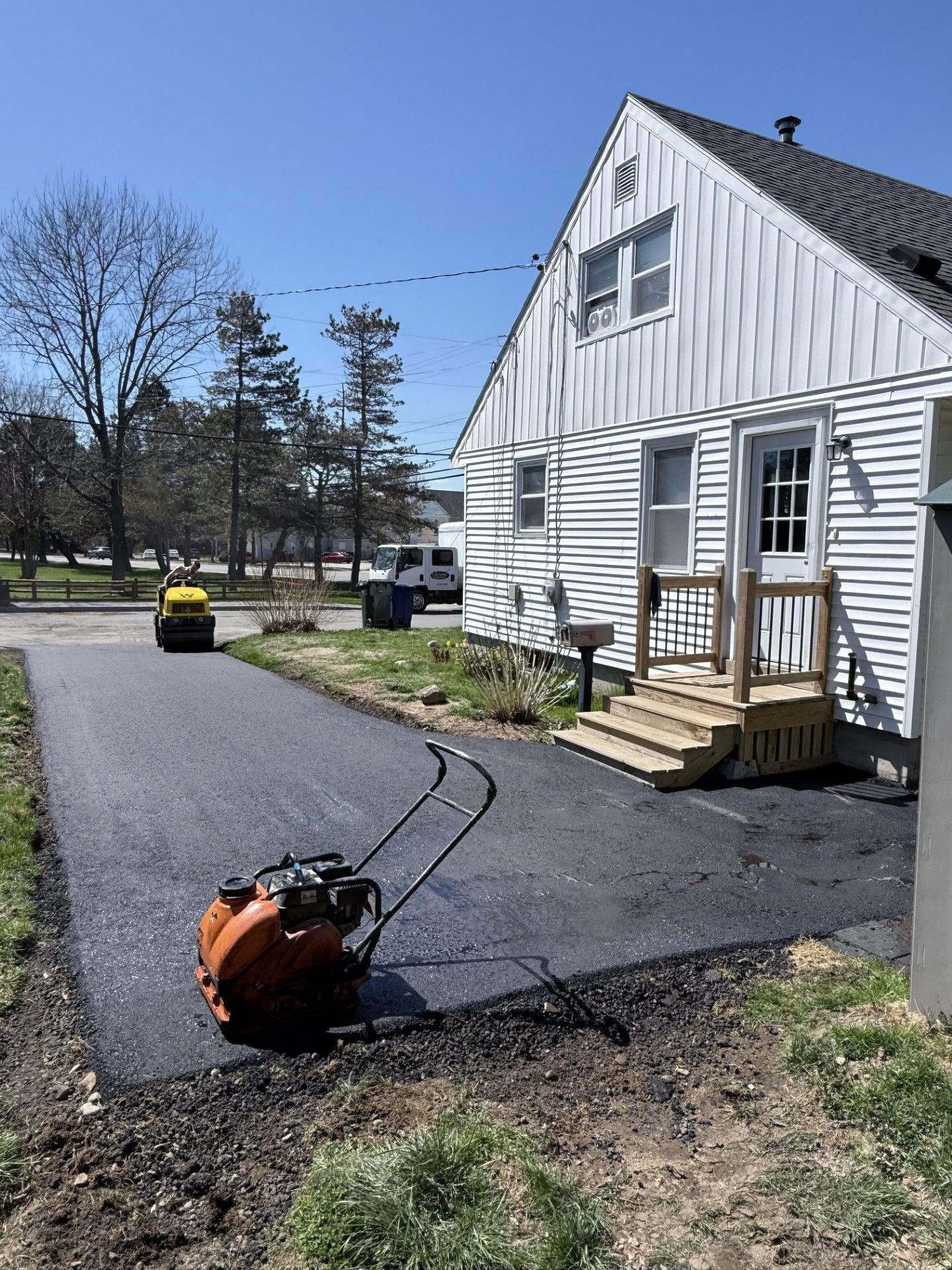 Fresh blacktop being laid beside a white house, with a worker in yellow and a paving machine in the driveway.