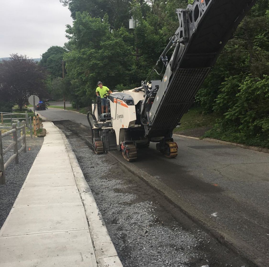 Road paving machine laying asphalt beside a sidewalk on a tree-lined street