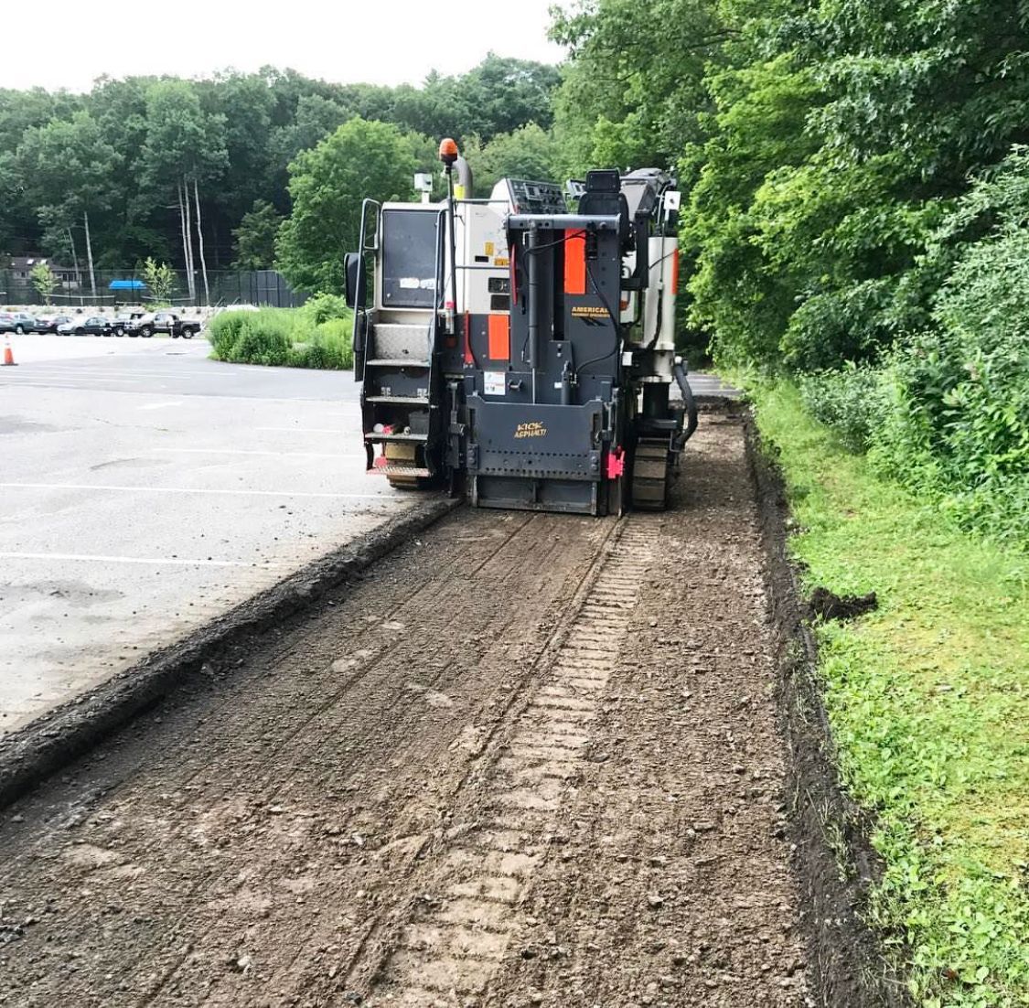 Road paver working along a muddy roadside next to a paved lane and green trees