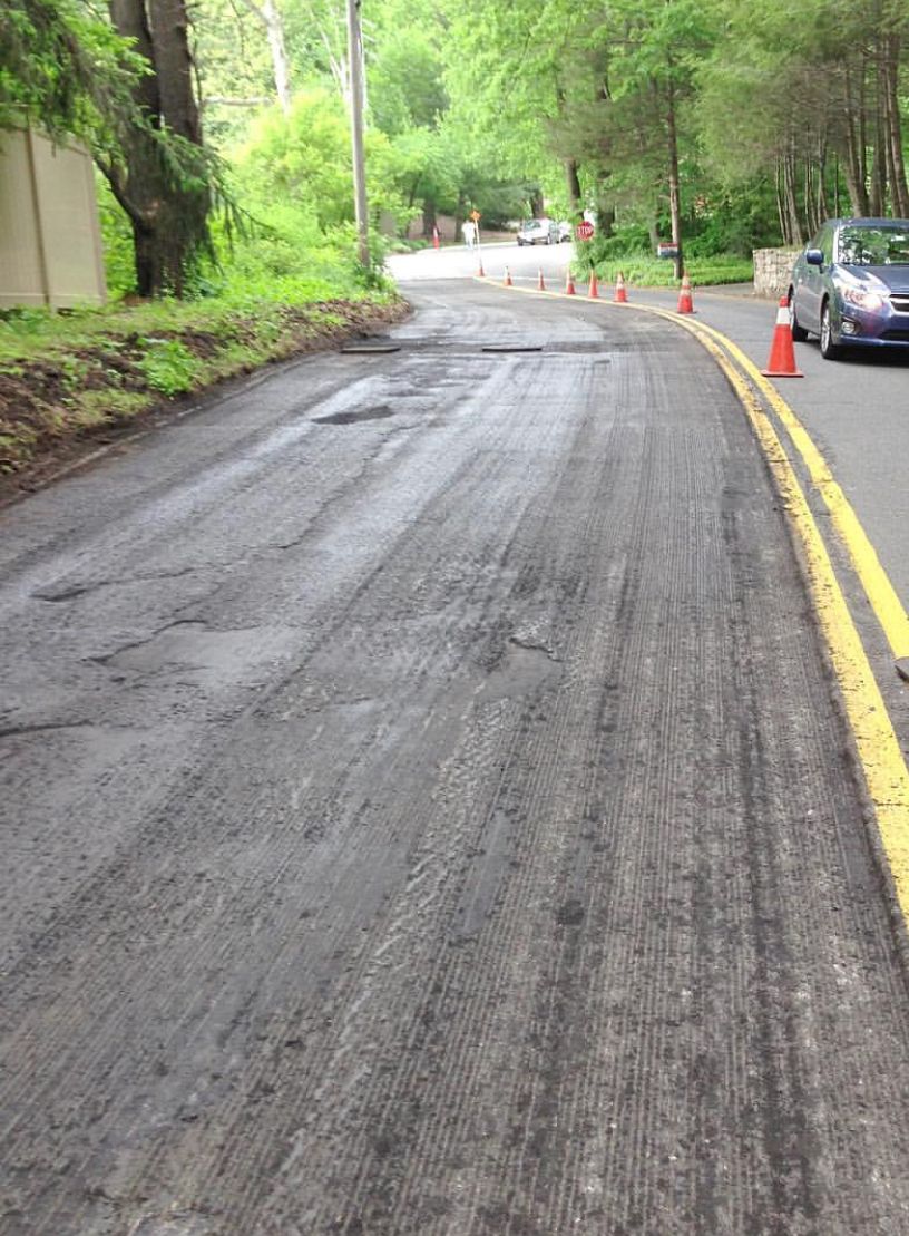 Wet road on a wooded hillside with orange traffic cones and parked cars along the curb