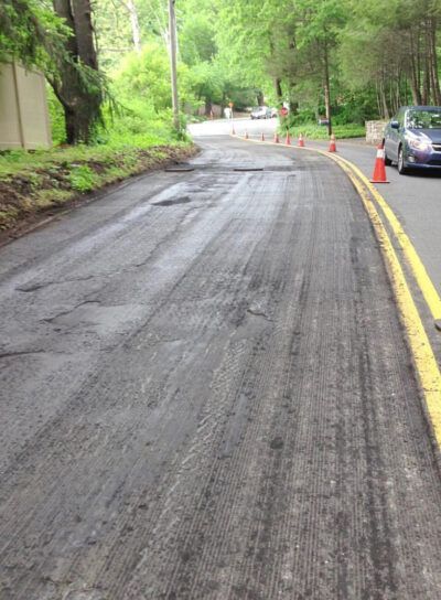 Wet winding road in a wooded area, with parked cars and orange cones along the curb