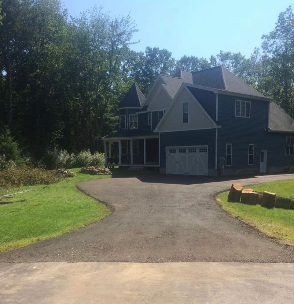 Dark gray house with a driveway, garage, and porch, surrounded by trees on a sunny day