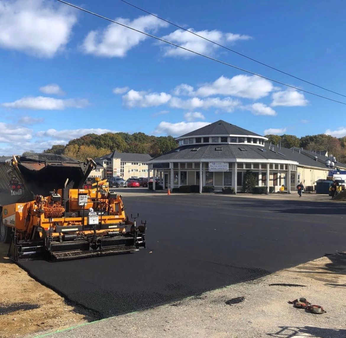 Road paving machine on fresh black asphalt in front of a white building under a blue sky