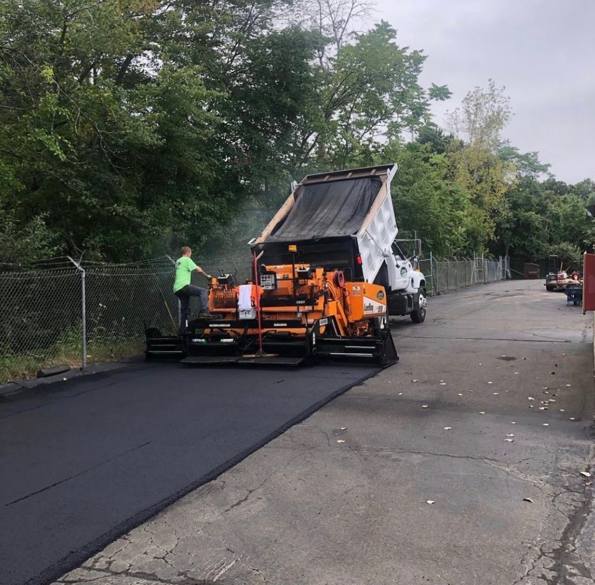 Orange paving machine laying fresh black asphalt on a road beside trees and a chain-link fence