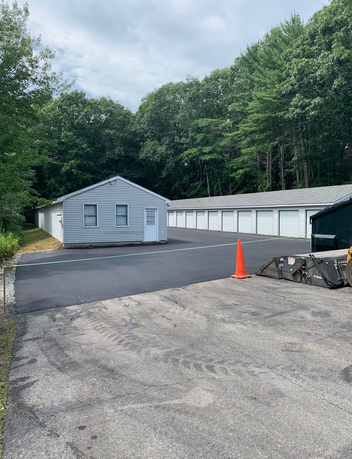 Gravel lot with white storage units, a small white building, trees, and an orange traffic cone