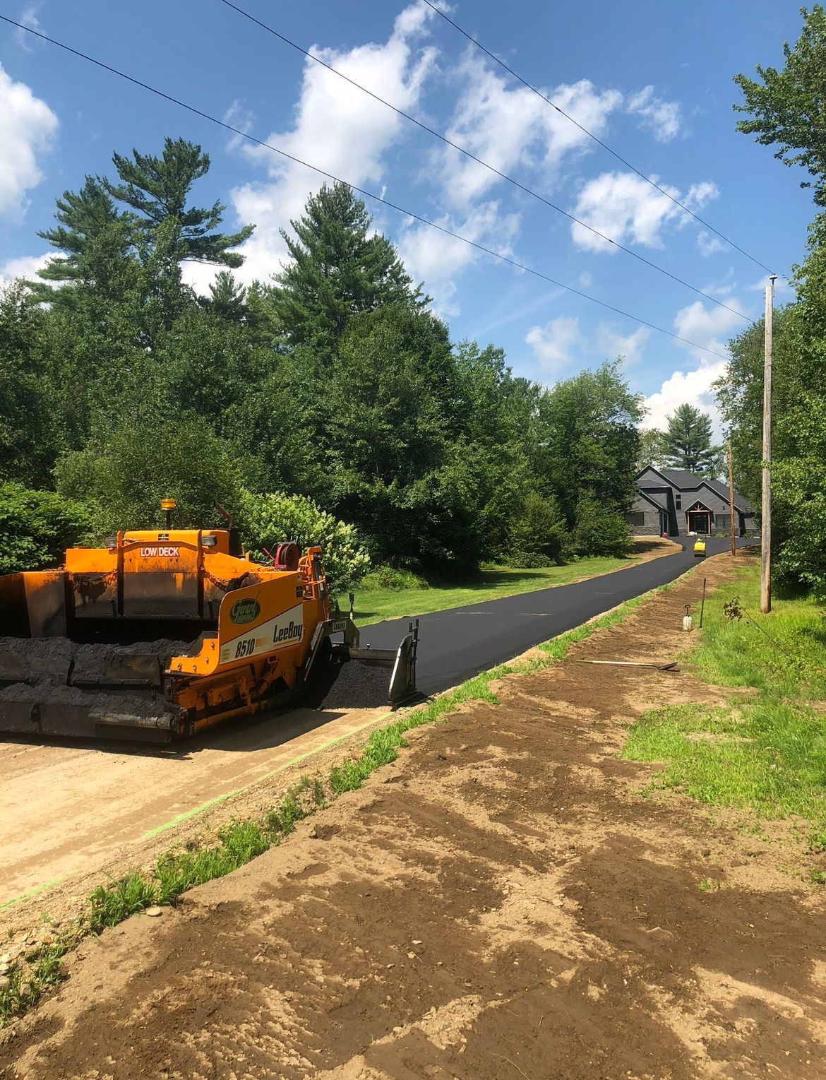Orange road paver laying black asphalt on a tree-lined path under a blue sky