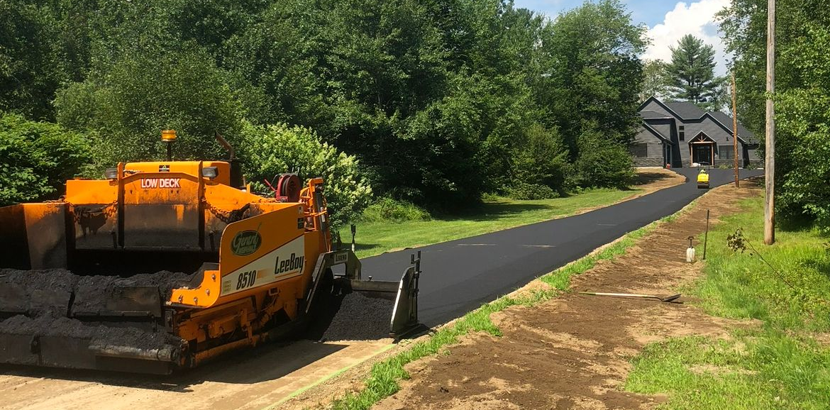 Orange paving machine laying fresh black asphalt on a road beside trees and a house