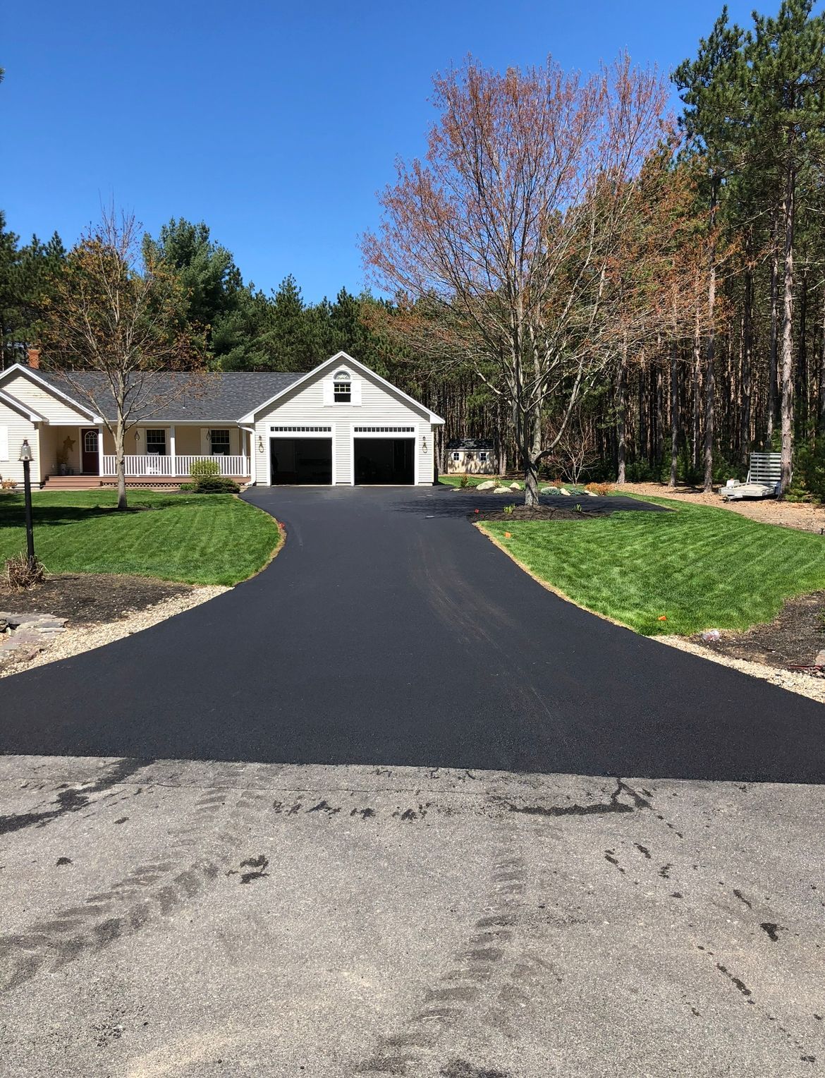 Suburban house with a newly paved black driveway, green lawn, and trees under a clear blue sky