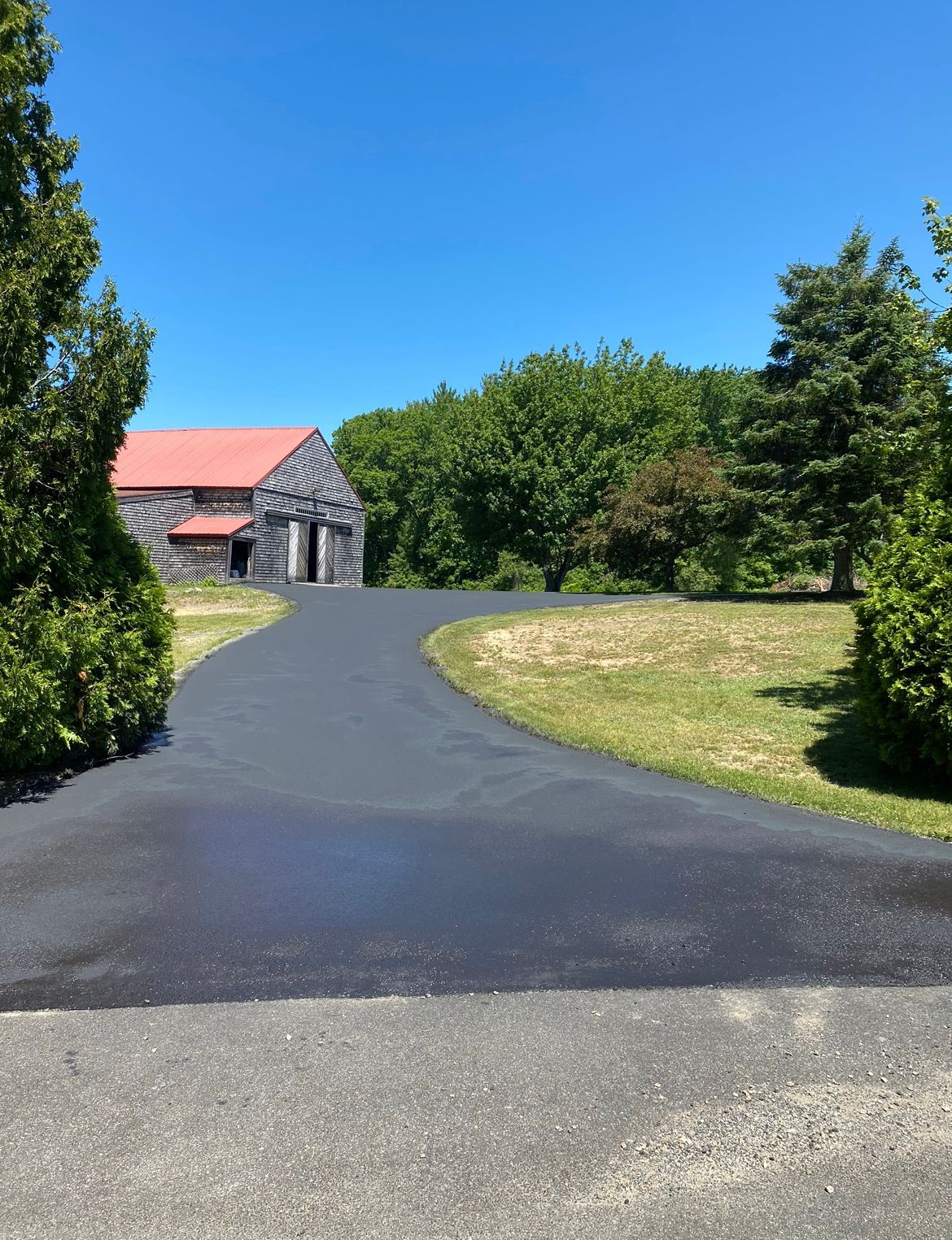 Curving paved driveway leading to a stone building with a red roof, surrounded by green trees under a blue sky