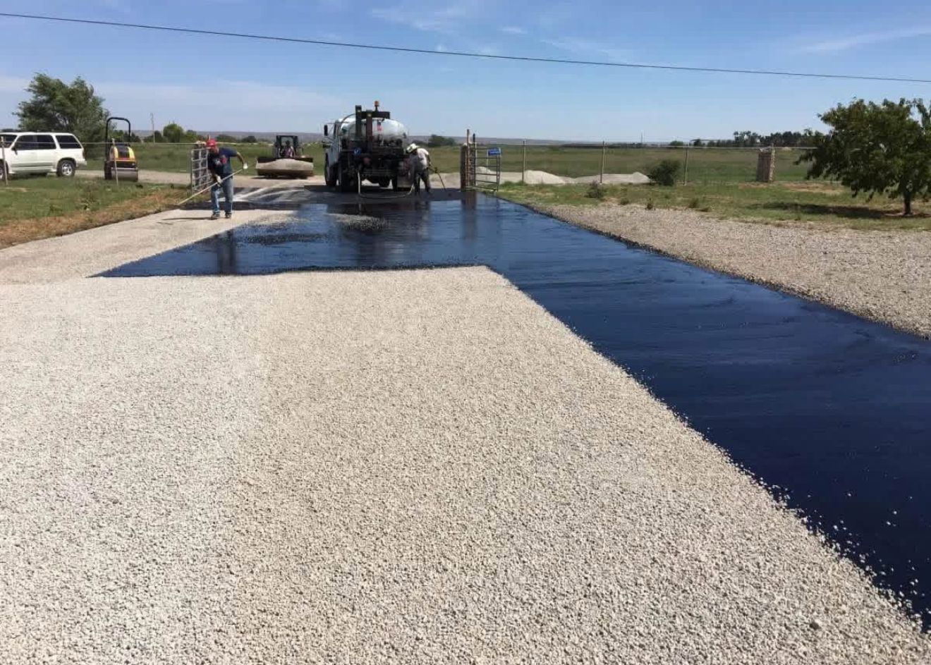 Road paving machine spreading dark asphalt on a gravel road beside parked vehicles.