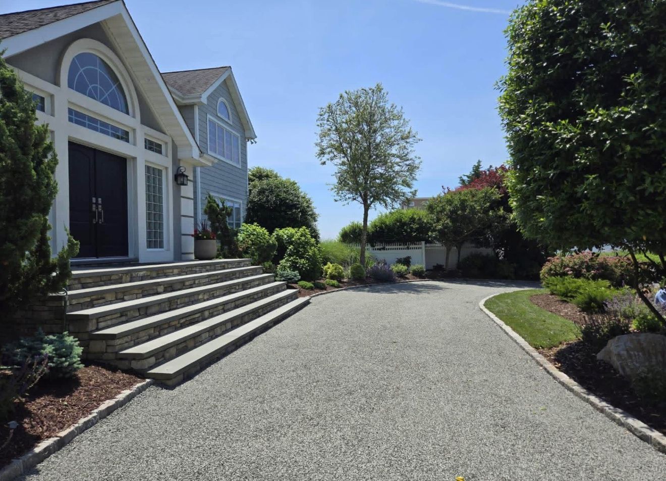 Driveway leading to a house with stone steps, arched entry, and landscaped garden under a blue sky