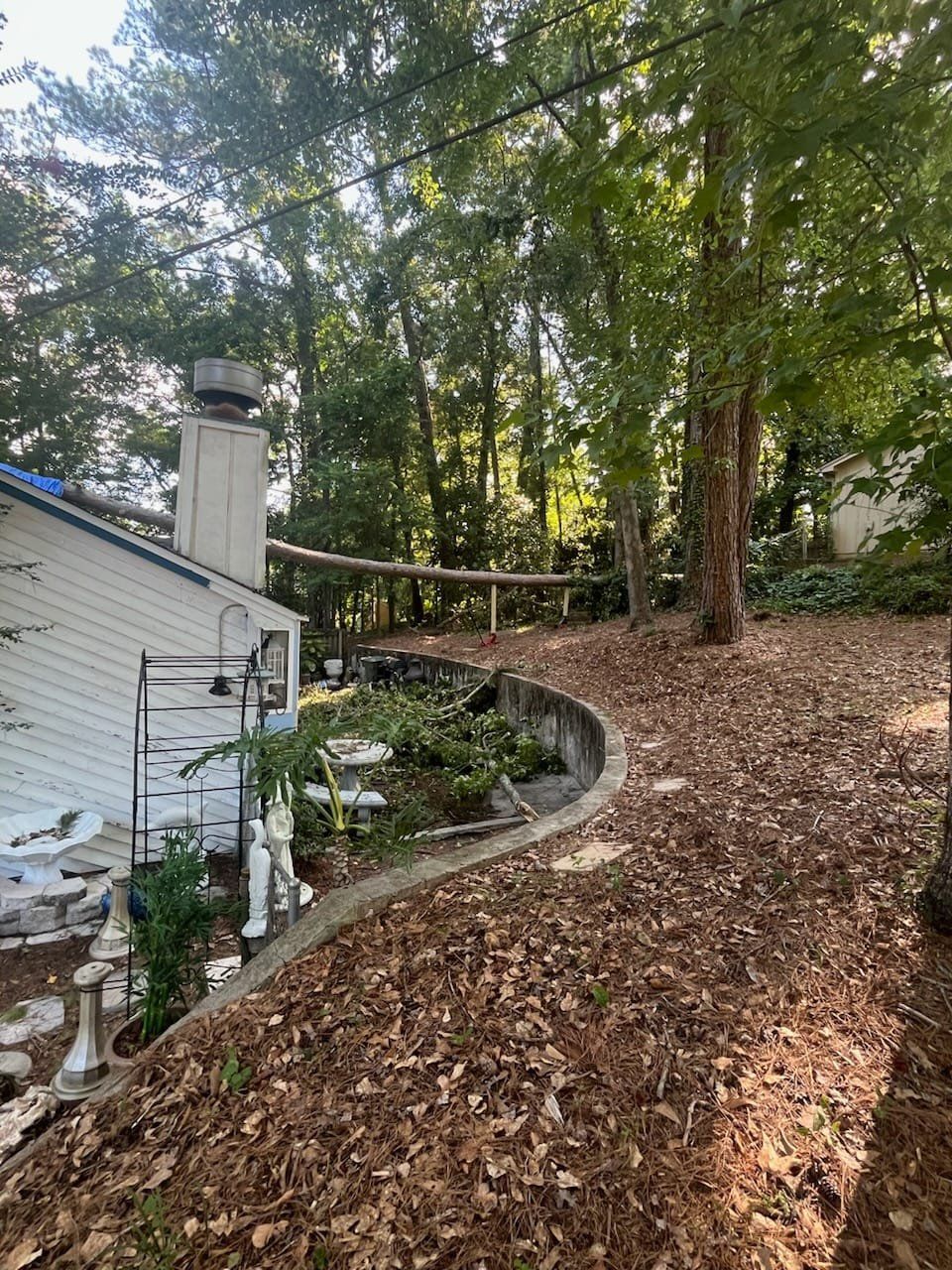 A backyard area features a curved concrete retaining wall, a white house with a chimney, and a heavily wooded lot.