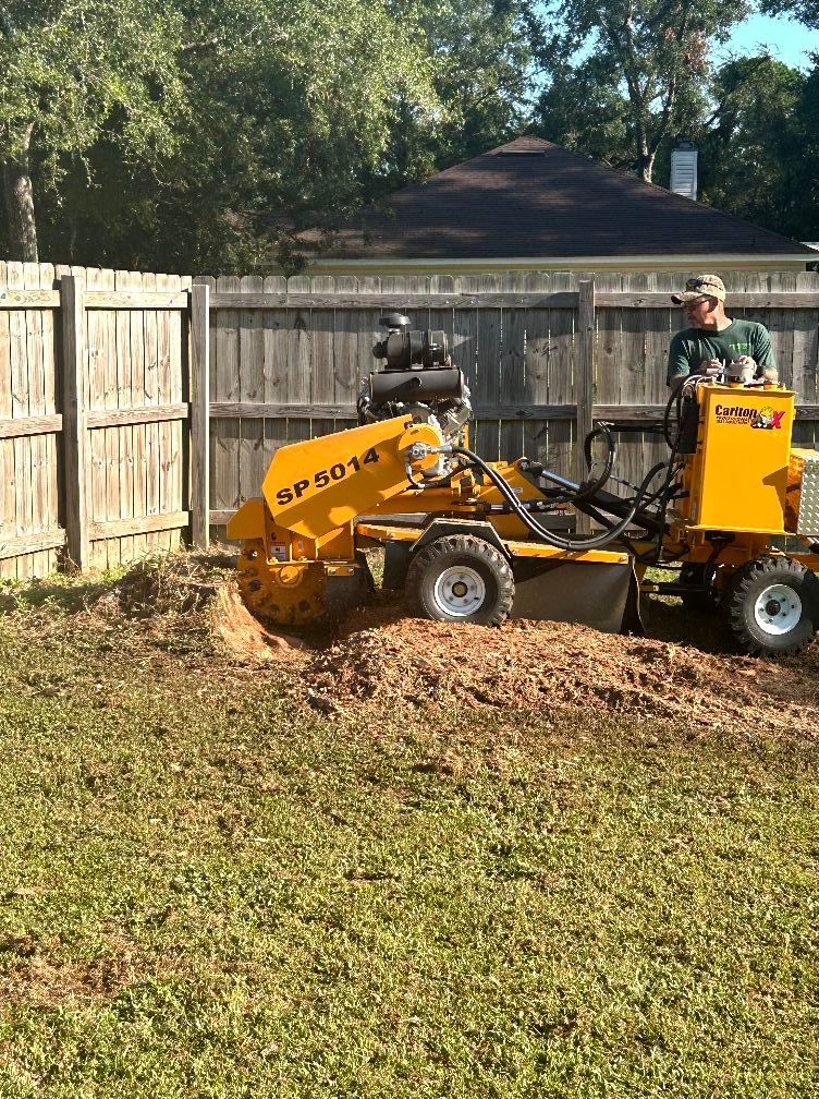A person operates a yellow SP5014 stump grinder on a grassy lawn in front of a wooden fence.