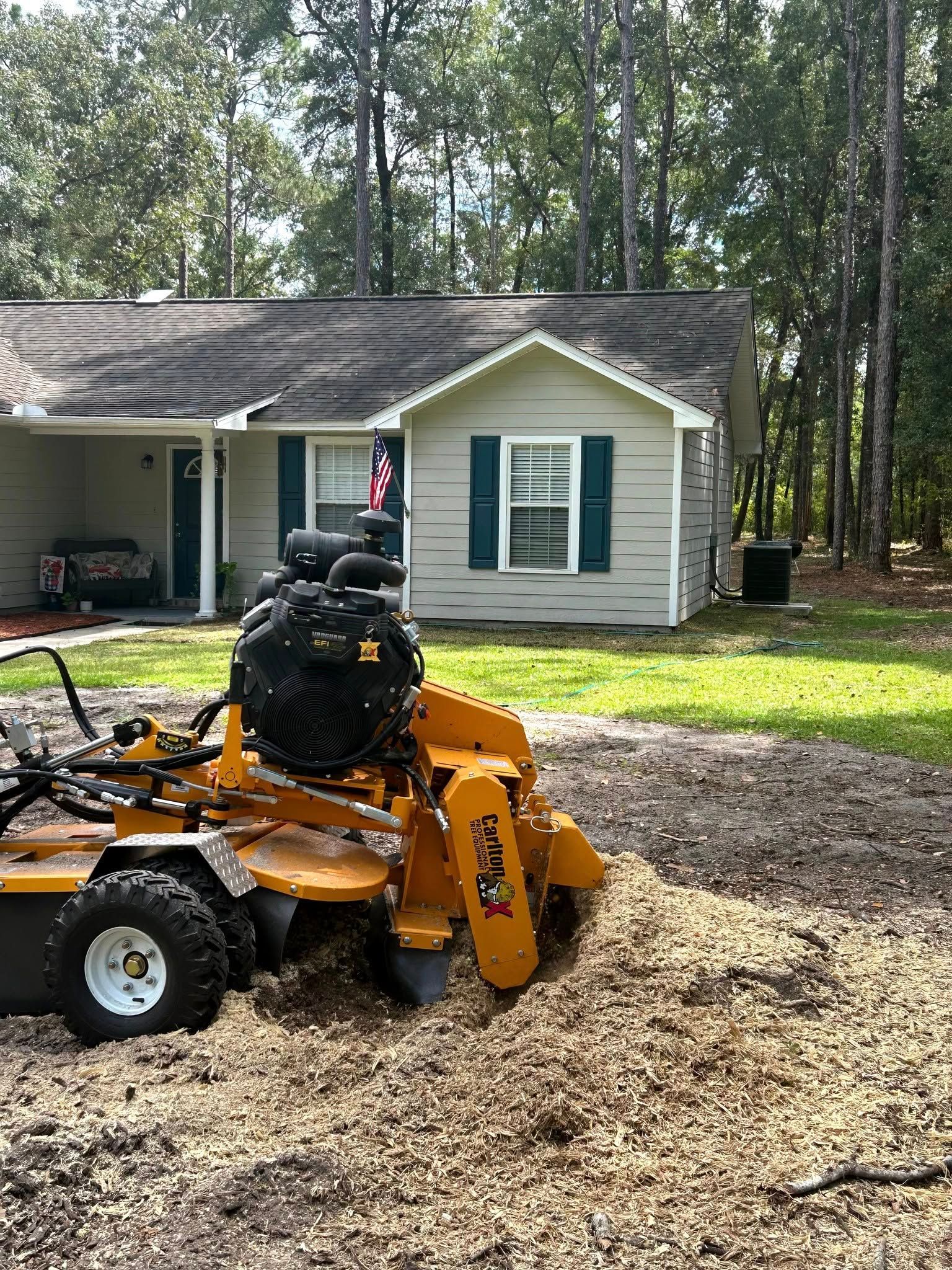 A yellow stump grinder sits on a pile of wood chips in front of a house with a beige exterior and green shutters.
