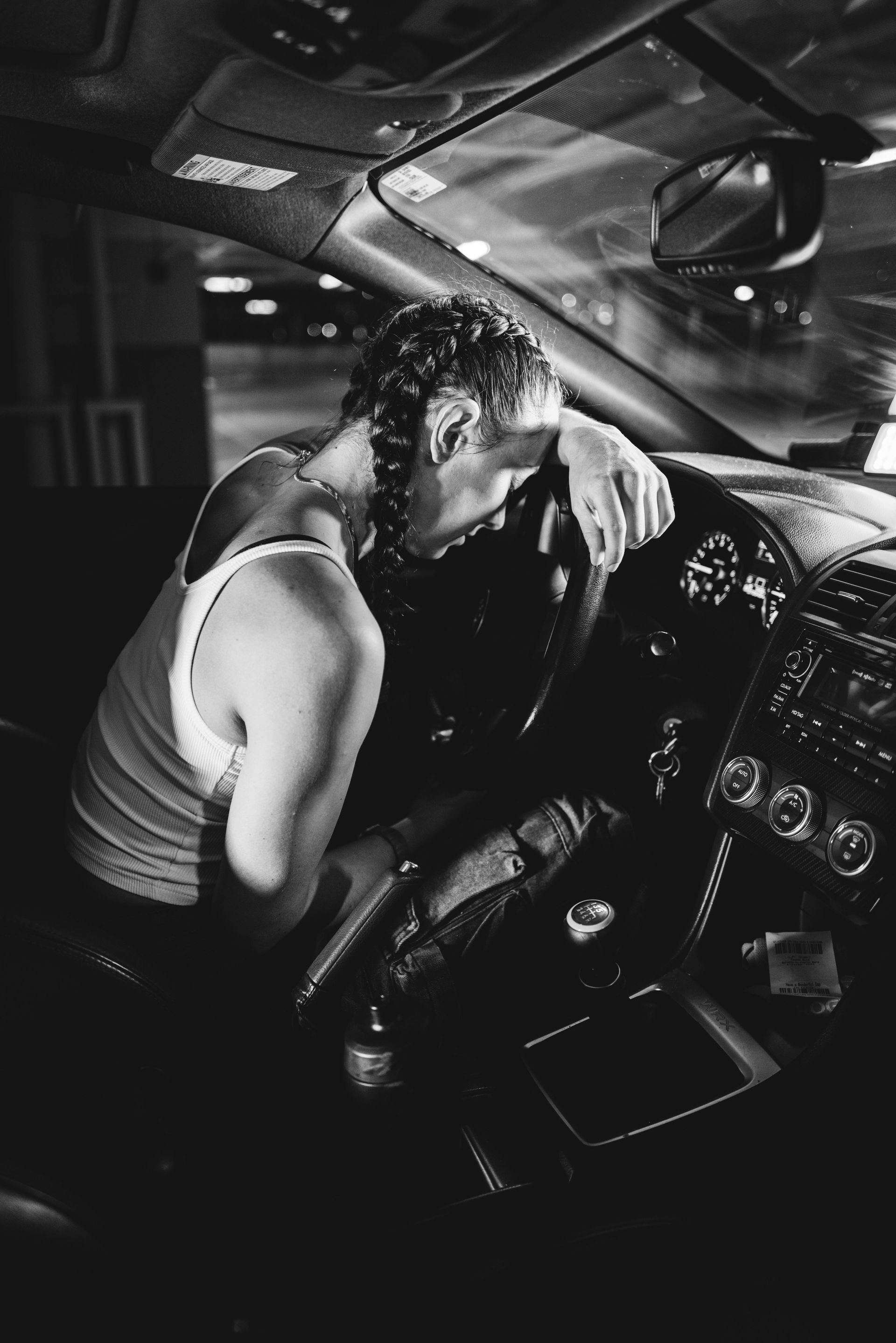 Person slumped over steering wheel inside car, appearing exhausted, in dark setting.