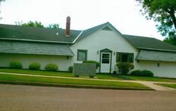 White one-story building with a green lawn, sidewalk, and a street. There is a sign in front.