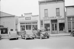 Black and white photo of a street in front of businesses with parked cars. A Coca-Cola sign is visible.