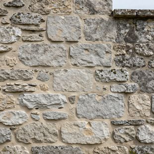A close-up view of a rustic stone wall built with light gray, irregularly shaped, weathered blocks and beige mortar.