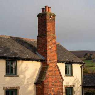 A brick chimney rises from the center of a rustic house with cream walls and a shingled roof under a soft sky.