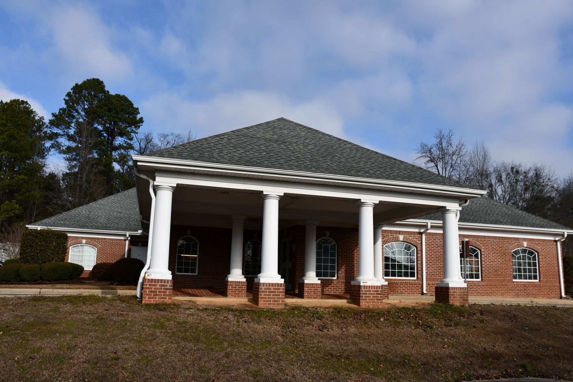 A brick building with a large covered entrance supported by white columns under a hip roof against a cloudy sky.