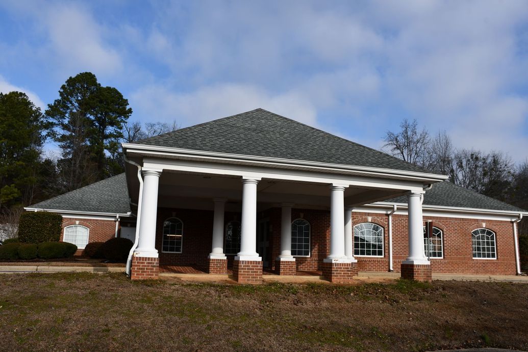 A brick building with a large covered entrance supported by white columns under a hip roof against a cloudy sky.