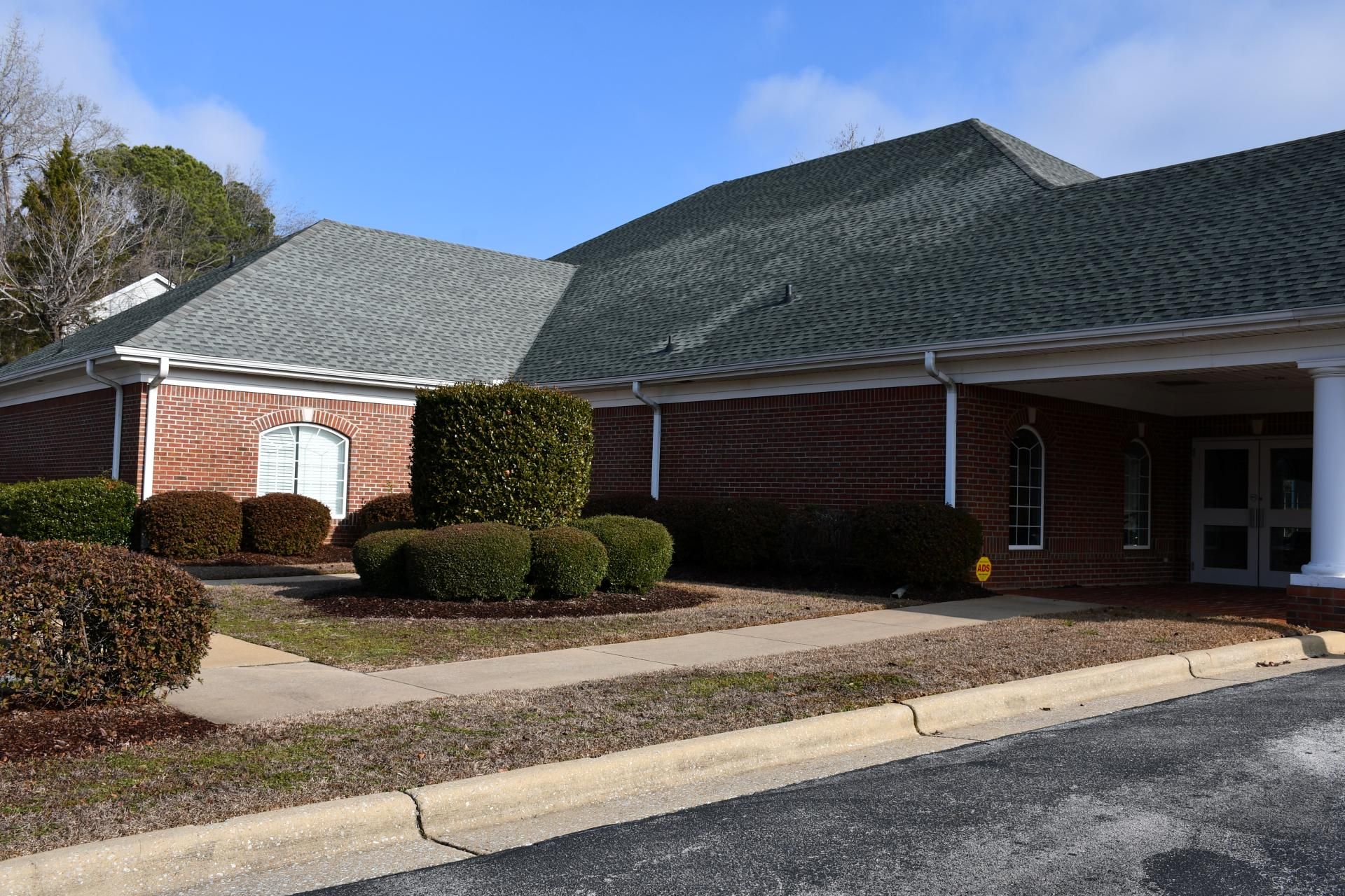 Low-angle view of a red brick building with a gray shingled roof, a covered entrance, and manicured shrubs in front.