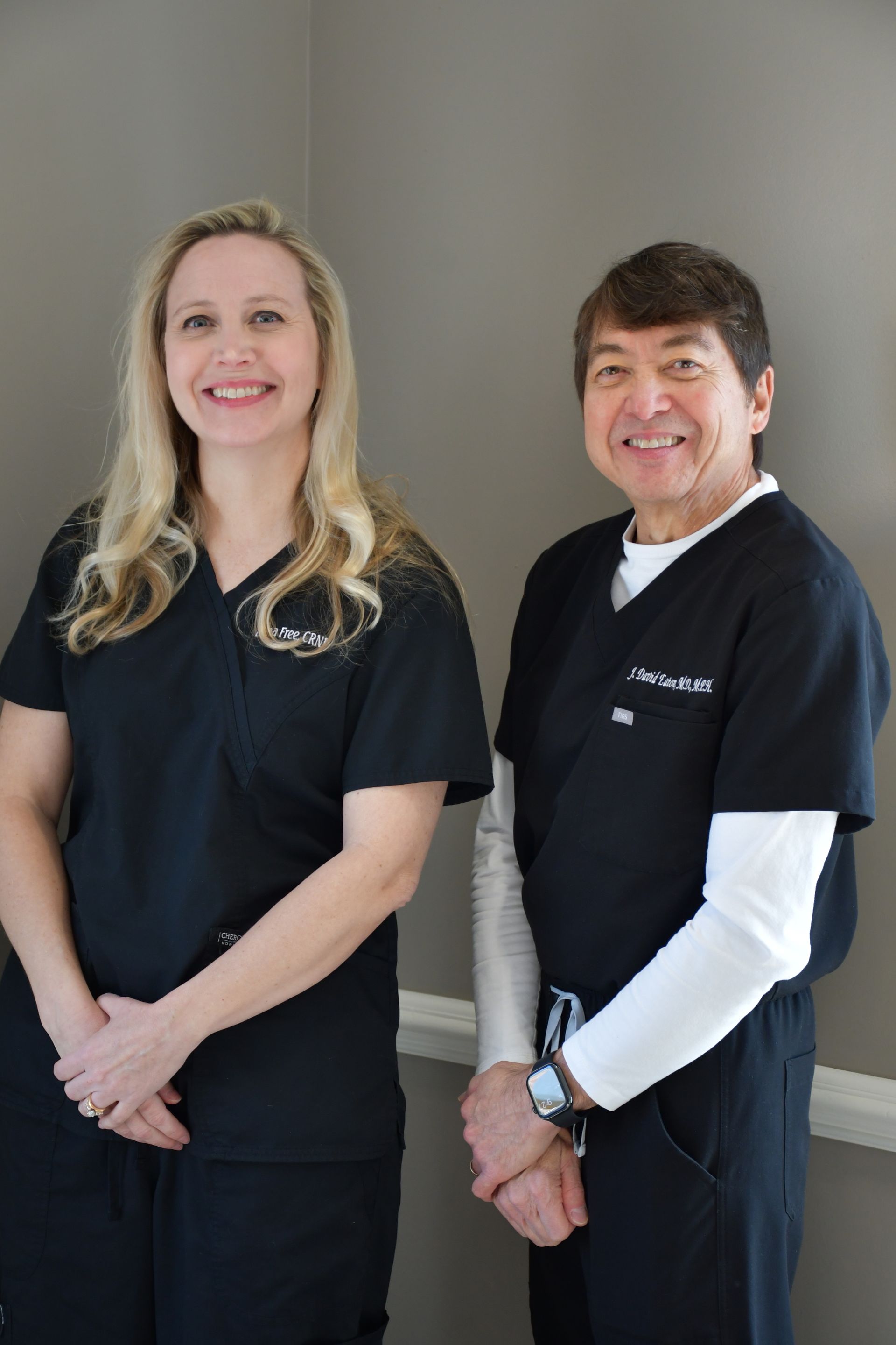 Two professionals in black scrubs smiling while standing side-by-side against a plain gray wall.