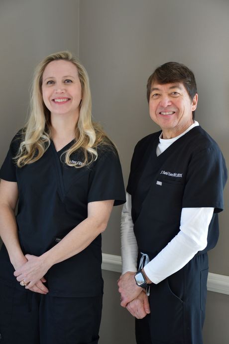 Two professionals in black scrubs smiling while standing side-by-side against a plain gray wall.