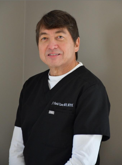 A person wearing a black scrub top over a white long-sleeved shirt standing against a plain gray wall.