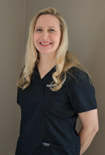 Smiling person with blonde hair wearing a dark blue scrub top against a plain gray wall.