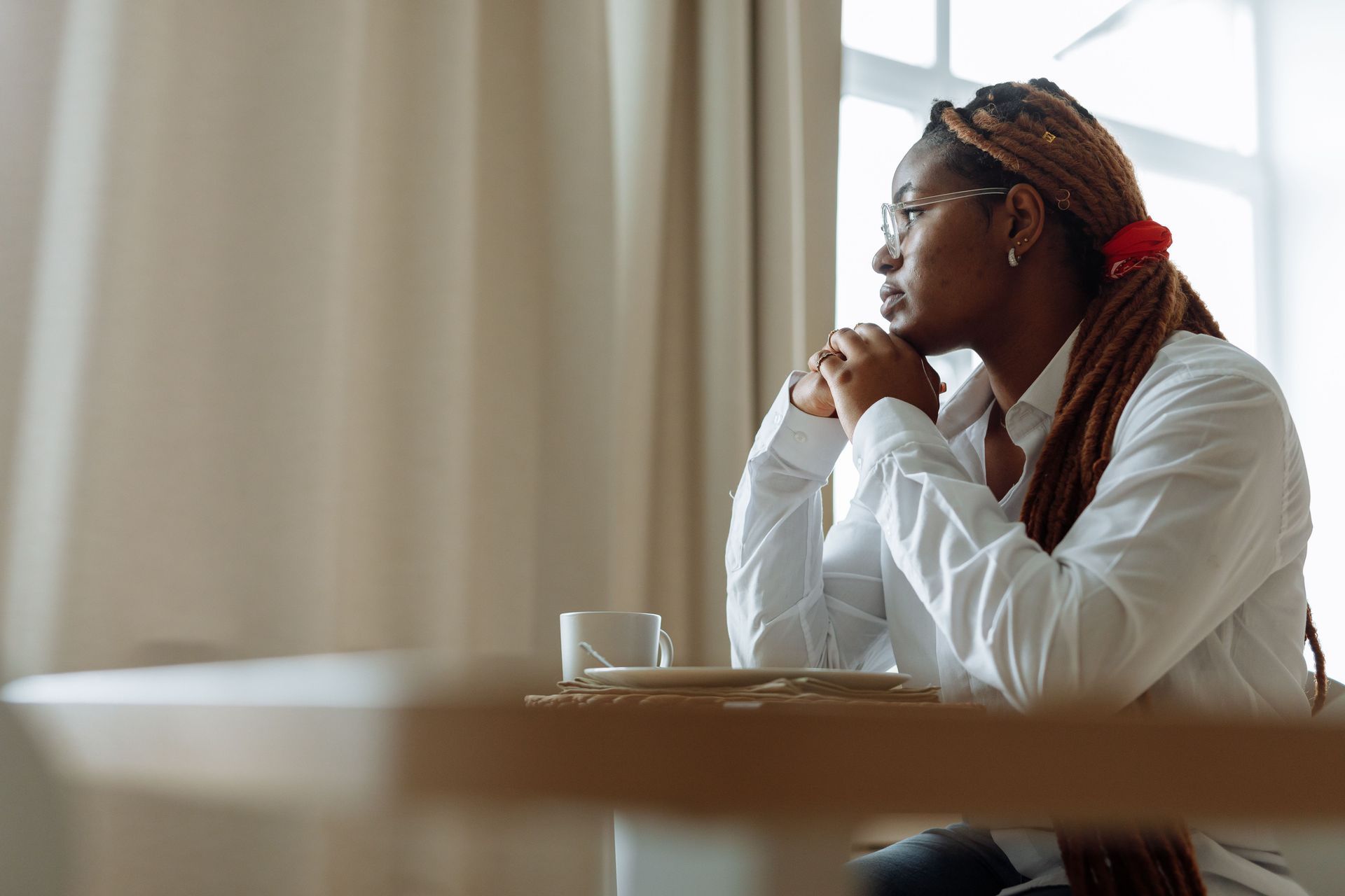 Person sitting thoughtfully at a table with hands clasped near a window
