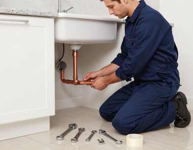 Plumber kneels under a sink tightening copper pipes with wrenches beside scattered tools.