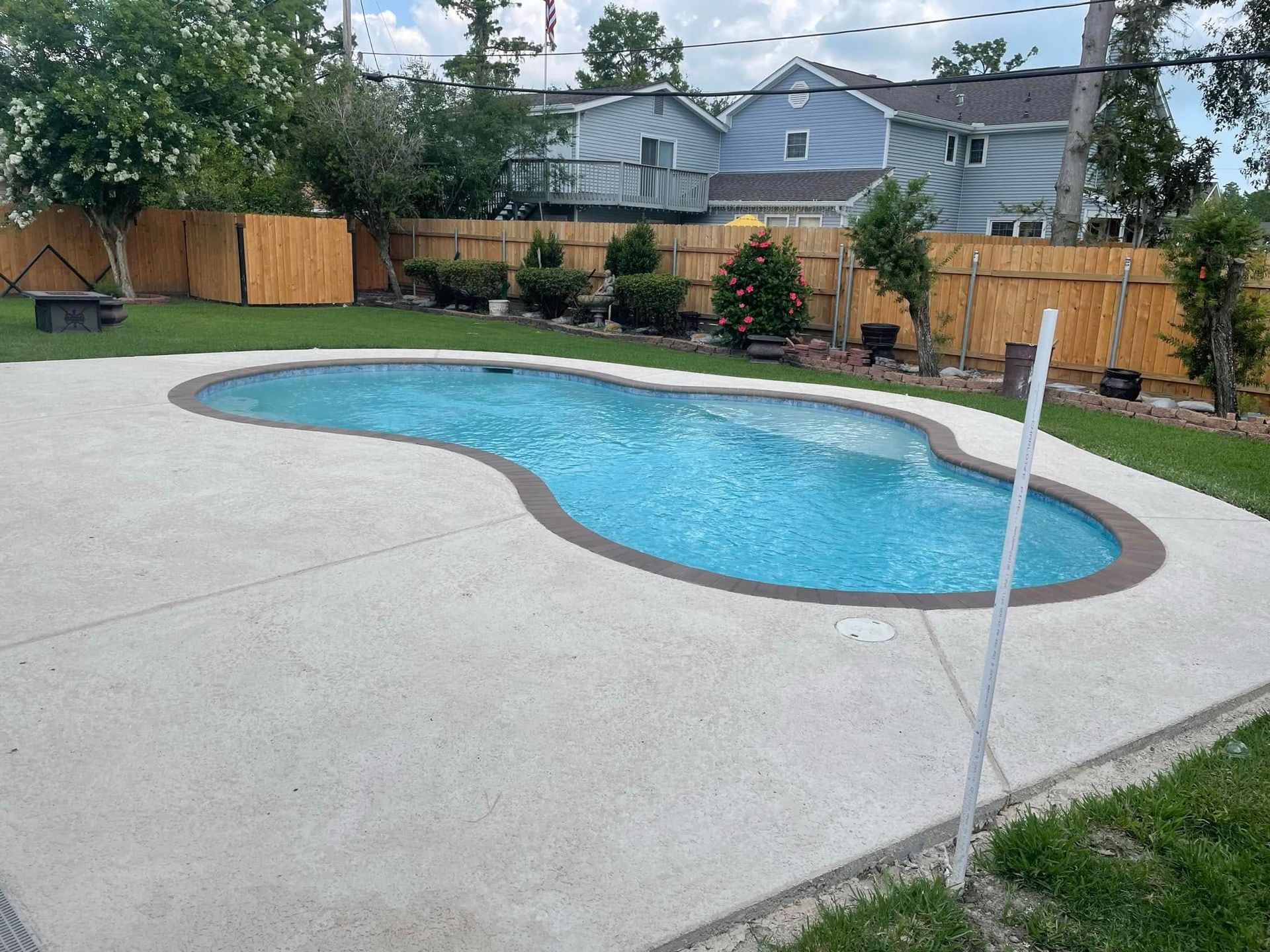 Backyard swimming pool with a concrete deck, wooden fence, and neighboring houses in the background